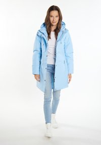 Lyseblå vandtæt parka lavet af syntetisk materiale, med hætte, lynlås og trykknapper, samt sidelommer, båret over jeans og sneakers.