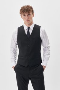 Junger Mann mit lockigem Haar, der eine schwarze Weste, ein weißes Hemd, eine schwarze Krawatte und schwarze Hosen trägt, stehen mit den Händen in den Taschen.