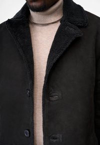Sort ruskindjakke med en struktureret, mørk pelskrave. Har to synlige knapper og bæres over en lys beige turtleneck sweater.