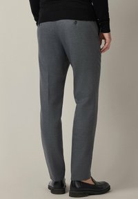 Szare spodnie na miarę o gładkiej fakturze i wąskim kroju, wyposażone w tylne kieszenie na guziki oraz czarne obuwie.