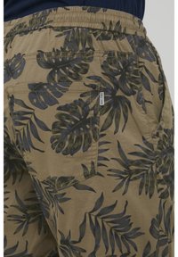 Pantaloni scurți khaki cu un model de frunze în verde închis și bleumarin. Au un costum elastic la talie și un buzunar pe spate cu un eticheta de marcă.