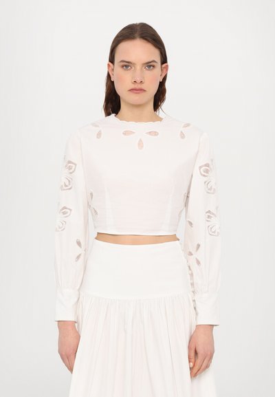 Blouse blanche coupée avec des manches longues, ornée de motifs floraux brodés et d'un décolleté festonné. Texture lisse et silhouette ajustée.