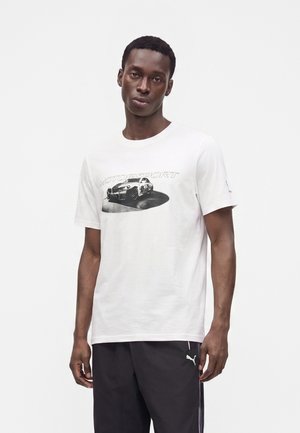 Homme portant un t-shirt blanc à manches courtes avec un graphisme noir et blanc d'une voiture de course et le mot "Motorsport,", associé à un pantalon de survêtement noir Puma.