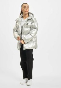 Sølv pufferjakke med hætte, der har en skinnende overflade og horisontalt quiltet mønster. Bæres over en hvid t-shirt og sorte bukser.