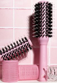 Deux brosses rondes roses avec des poils noirs, des rouleaux de cheveux texturés roses et une pince à cheveux transparente sur un fond en carreaux roses.