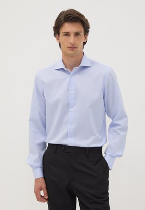 Junger Mann, der ein hellblau gestreiftes Langarmhemd und schwarze Hose trägt, steht vor einem schlichten weißen Hintergrund.