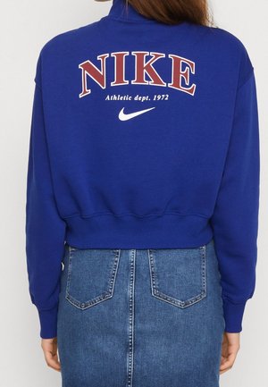 Osoba ubrana w niebieską, krótką bluzę z napisem „NIKE Athletic dept. 1972” i białym logo Swoosh na plecach, w połączeniu z wysokim stanem spódnicy jeansowej.