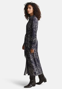 Μέχρι το μέσο της γάμπας φόρεμα σε ναυτικό μπλε με λουλουδάτα σχέδια, μακριά μανίκια, γιακά και ζώνη στη μέση· διαθέτει χρυσά λουλουδάτα μοτίβα και πλαϊνή σκίσιμο, συνοδευόμενο από καφέ μποτάκια.