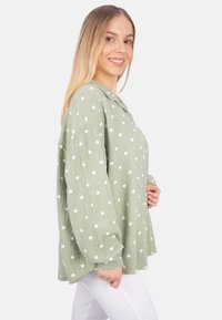 Green, textured shirt with a button-up collar, featuring a white heart pattern, and long sleeves, worn over white pants.