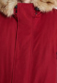 Röd parka-jacka med en krage av konstpäls, dragkedjestängning och en slät, matt yta med sidofickor och förstärkt sömnad.