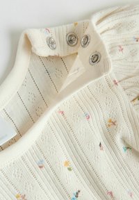 Tissu en tricot crème avec de petits motifs floraux multicolores et trois boutons-pression argentés sur l'encolure d'un vêtement.