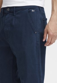 Tume sinised püksid, millel on kitsas lõige, esipocketi disain, metallist nööbiga sulgemine ja väike brändielement tasku kohal.