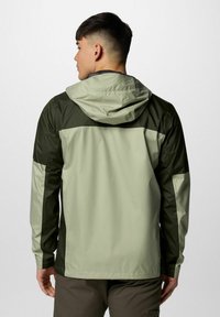 Mężczyzna odwrócony plecami, ubrany w dwukolorową, jasną i ciemnozieloną kurtkę z kapturem i długimi rękawami, oraz ciemne spodnie.