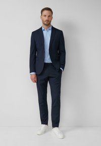 Marineblaues Anzug mit einem maßgeschneiderten Blazer, passenden Hosen und einem hellblauen Polka-Dot-Hemd. Weiße Sneaker runden das Ensemble ab.