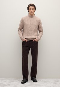 Maglione beige a maglia cable con colletto alto, abbinato a pantaloni dritti marrone scuro e mocassini neri, su uno sfondo neutro.