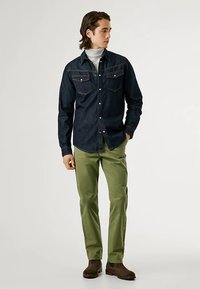 Camisa escura de ganga com botões e bolsos no peito, combinada com calças verdes justas e botins castanhos até ao tornozelo. Gola alta macia por baixo.