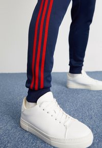 En person som bär marinblå byxor med röda ränder och vita sneakers, stående på ett blått mattlagt golv.