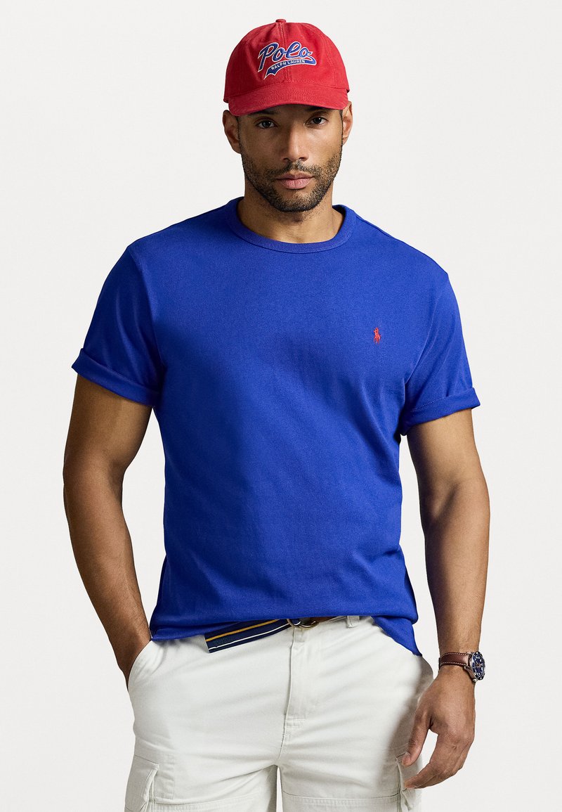 Βασιλικό μπλε κοντομάνικο μπλουζάκι με ρολάρισμα στις μανσέτες και ένα μικρό κόκκινο κεντητό λογότυπο στο στήθος, συνδυασμένο με ανοιχτόχρωμες cargo βερμούδες.