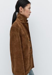 Brązowa zamszowa kurtka z wysokim kołnierzem, zapięciem na guziki i kieszeniami po bokach. Gładka faktura i dopasowany krój, pokazana z boku.