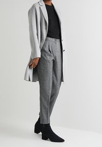 Cappotto di lana grigio chiaro, top a pois neri, pantaloni grigi su misura e stivali neri alla caviglia. Texture lisce, taglio classico e dettagli con bottoni.