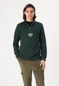 Mørkegrønn zip-up hettegenser med stående krage, prydet med hvit "BOSTON" tekst. Kombinert med ribbede olivengrønne cargo bukser.