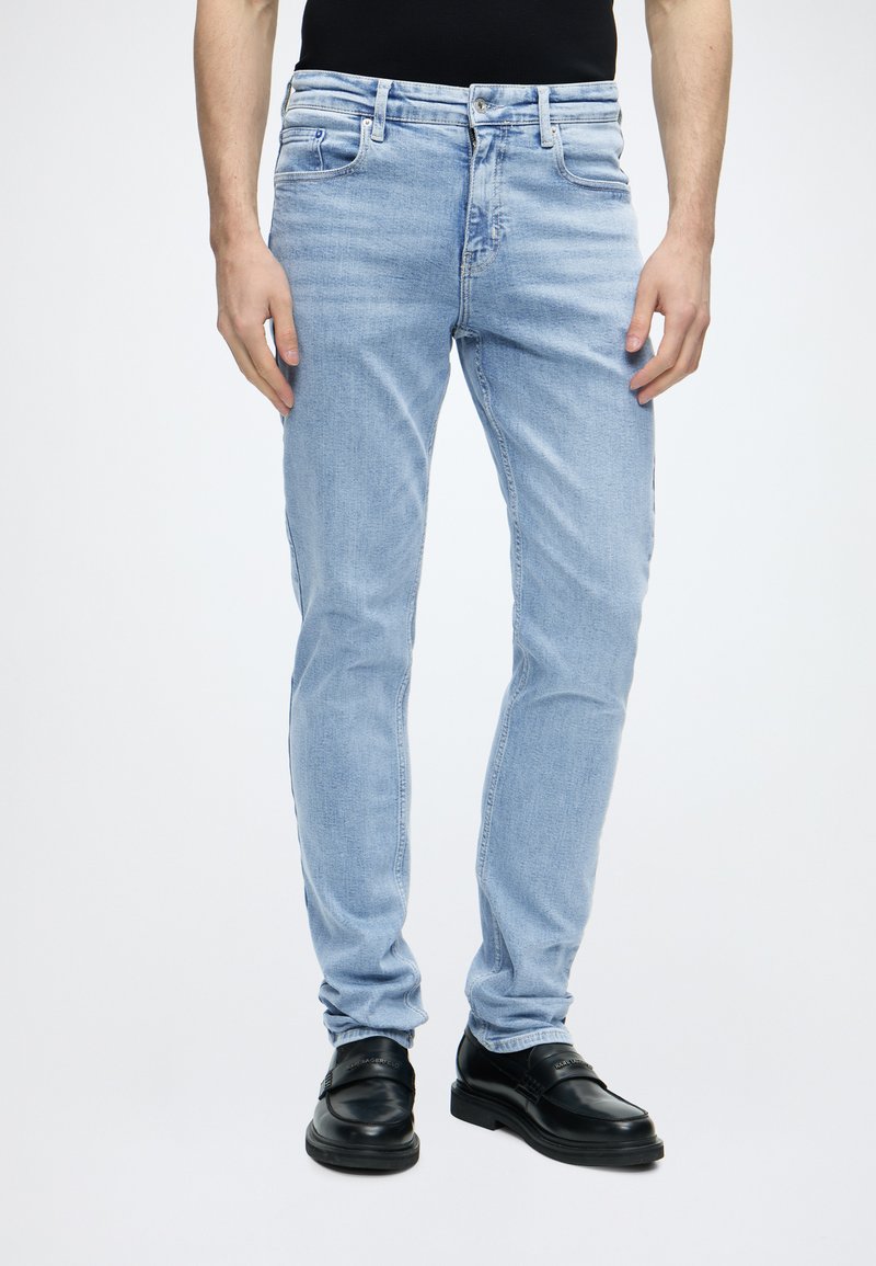 Τζιν slim fit ανοιχτού μπλε χρώματος που φοριέται από ένα άτομο που στέκεται, συνδυασμένο με μαύρα δερμάτινα loafers και μαύρο τοπ, σε λευκό φόντο.