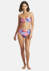 Kobieta stojąca boso, ubrana w strapless, kolorowy kostium kąpielowy w abstrakcyjny wzór z wysokim stanem na białym tle.