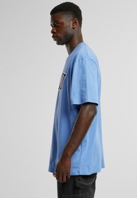 Tricou din bumbac de culoare albastră deschisă, cu mâneci scurte, gât rotund și imprimeu cu litere negre pe față. Țesătura pare moale și ușor texturată.