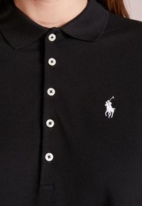 Schwarzes Poloshirt aus strukturiertem Stoff, mit klassischem Kragen, fünf weißen Knöpfen und einem kleinen weißen gestickten Logo.