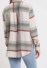 Long-sleeve shirt featuring a light beige fabric with black and rust-colored striped patterns, and a rounded hem. Buttoned cuffs are visible.