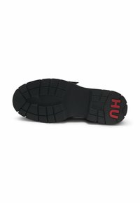 Semelle de chaussure en caoutchouc noir avec des motifs antidérapants texturés et le logo rouge "HU" près du talon. Présente une forme large et plate pour la stabilité.