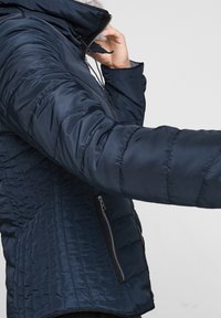 Muž v námořnickém prošívaném bundě s zipovými kapsami, upravující límec u brady na světlém jednobarevném pozadí.