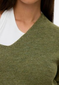 Close-up van een persoon die een witte geribde tanktop onder een groene gebreide V-hals trui draagt, waarbij het nek- en schoudergebied zichtbaar is.