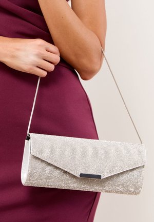 Frau in burgunderrotem Kleid hält eine silberne, funkelnde Clutch mit dünnem Metall-Schulterriemen vor einfachem Hintergrund.