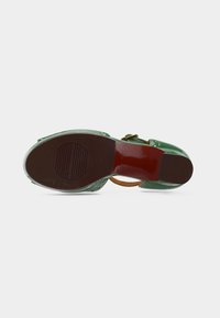 Chaussure en cuir verni vert avec une bride à la cheville, dotée d'une semelle en caoutchouc marron et d'un détail texturé. Semelle plate avec une légère courbure.