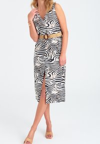 Černobílé šaty se zebrovaným vzorem bez rukávů, s výstřihem do V a bočním rozparkem, které jsou staženy béžovým pleteným opaskem. Žena má na sobě béžové sandály.