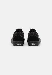 Un paio di sneakers Vans nere viste dal tallone, con cuciture bianche e loghi "Off The Wall" sulla suola in gomma nera.
