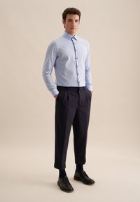 Camicia button-up azzurro chiaro con colletto, pantaloni scuri a pieghe, scarpe nere e calzini; lo sfondo semplice mette in risalto il design degli indumenti.