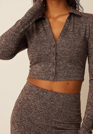 Kvinde iført en brun meleret strikket cropped skjorte med knapper og lange ærmer samt et matchende højtaljet nederdel.