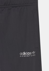 Černé sportovní kalhoty s hladkou texturou, které mají elastický pas a boční kapsy. Na noze je vytištěno stříbrné logo "adidas ADVENTURE".