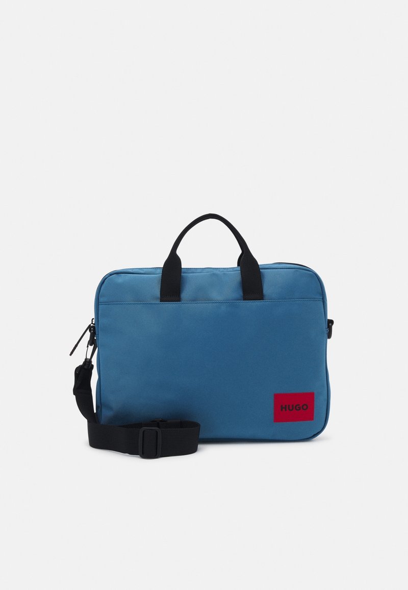 Serviette rectangulaire bleue avec poignées noires et bandoulière réglable, avec étiquette rouge "HUGO" en bas à droite à l'avant.