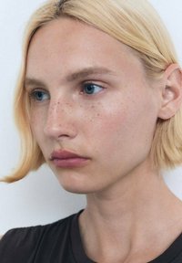 Nahaufnahme einer Person mit kurzem blonden Haar und heller Haut, die blaue Augen und sichtbare Sommersprossen hat, und ein dunkles Oberteil vor einem hellen Hintergrund trägt.