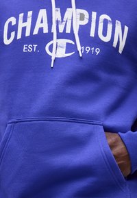 Μπλε φούτερ με κουκούλα με λευκή επιγραφή "CHAMPION EST. 1919" και λογότυπο, λευκά κορδόνια και το χέρι ενός ατόμου εν μέρει στην μπροστινή τσέπη.