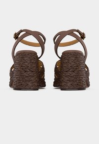 Καφέ πλεκτές σανδάλες με χοντρό τακούνι και λουράκια με αγκράφα στον αστράγαλο, εμφανιζόμενες από πίσω σε λευκό φόντο.