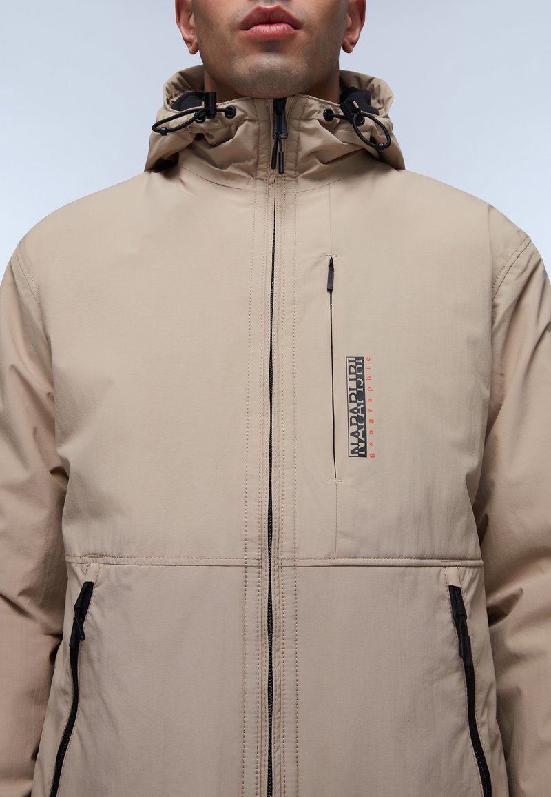 Beige waterdichte jas met een hoge kraag, verstelbare capuchon en zwarte ritsen. Beschikt over een ritszak op de borst en logo-detail.