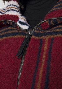 Veste en polaire rouge avec un col haut, présentant un motif rayé en bleu, marron et blanc, et une fermeture éclair noire avec des éléments métalliques.