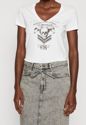 Persoon met een witte V-hals T-shirt met schedel- en arendgrafiek en tekst, gecombineerd met een hooggetailleerde grijze denim rok met strikceintuur.