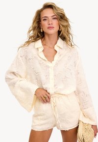 Femme avec des cheveux blonds ondulés portant une chemise boutonnée texturée crème et un short assorti, tenant une pochette en paille tissée dans sa main gauche.