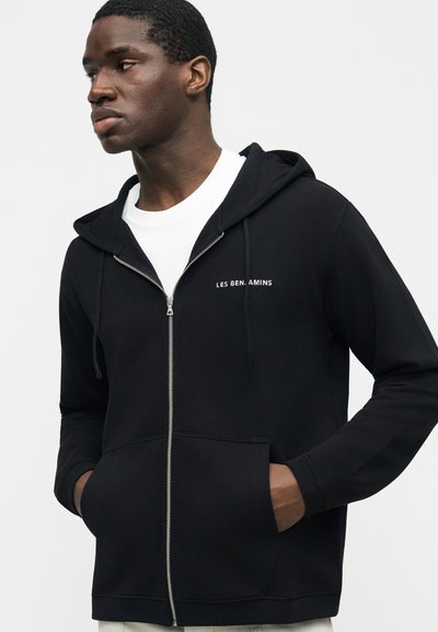 Άνδρας που φοράει μαύρο φούτερ με φερμουάρ και λευκό κείμενο στο στήθος, στέκεται με τα χέρια στις τσέπες, κοιτάζοντας προς τα πλάγια μπροστά σε απλό φόντο.