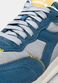 Blue suede sneaker with grey textile panels, flat grey laces, yellow stitching accents, and a white rubber sole. Standard shape and design.